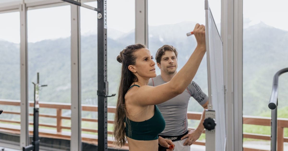 Whiteboard in a gym showing a workout program with set counts being crossed out and rewritten