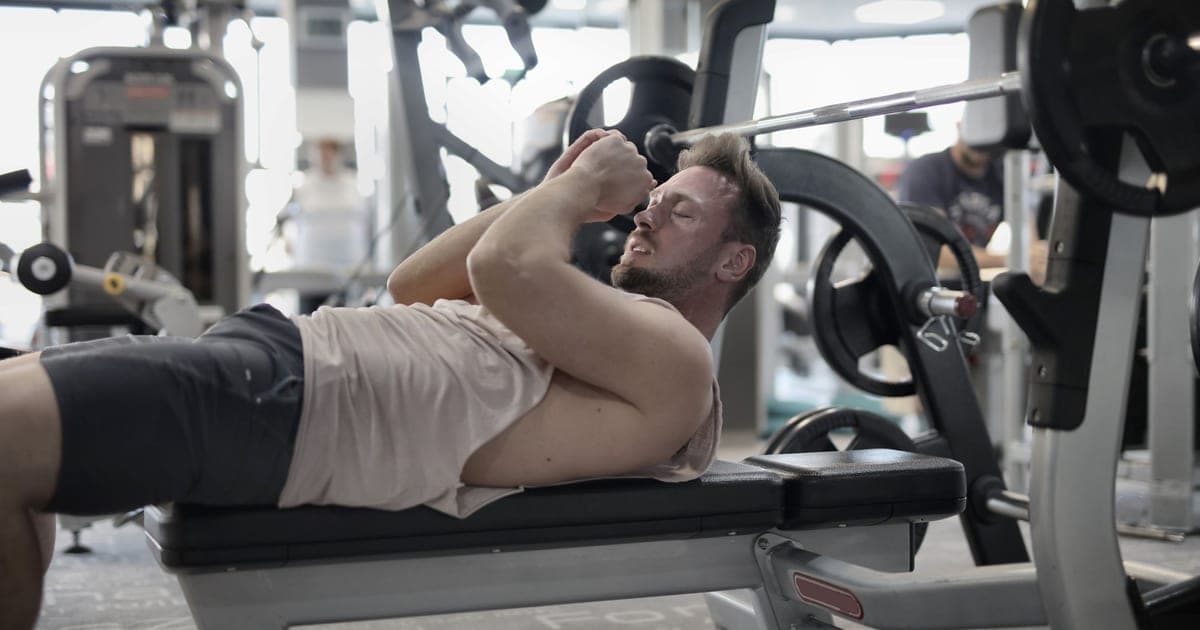 Lifter sitting on a bench between sets with a gym wall clock behind him, illustrating rest between sets under time pressure