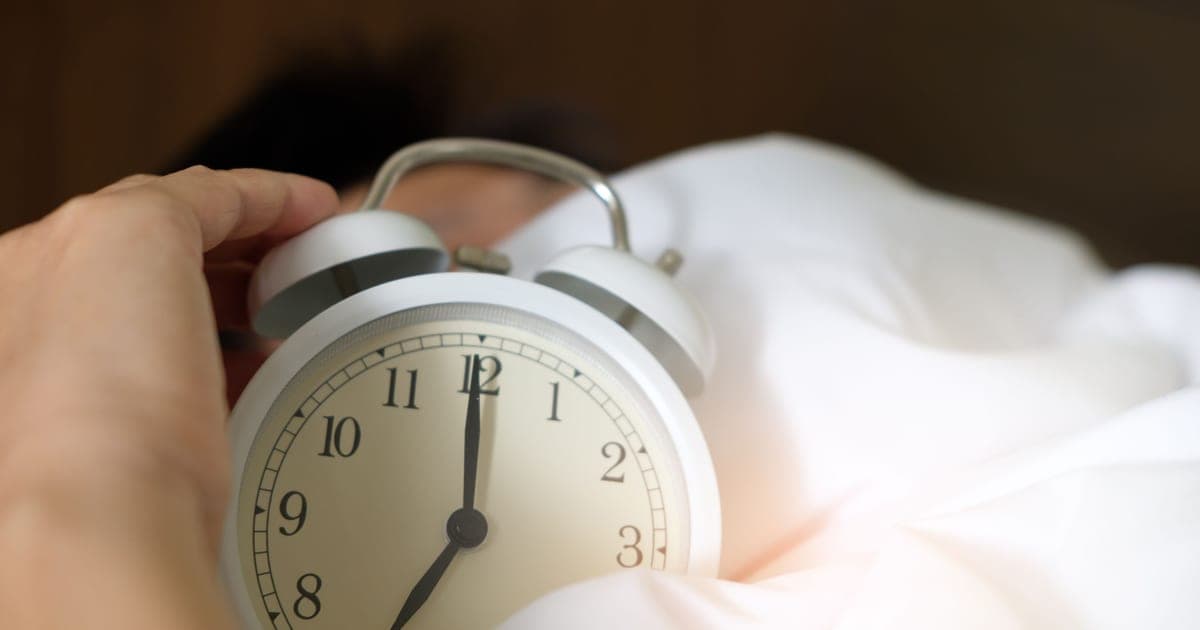 Person sitting on the edge of a bed in a dark room with an alarm clock showing early morning, looking tired before a training session