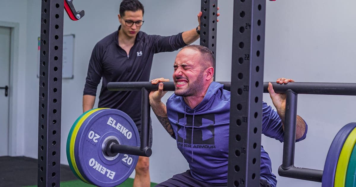Older professional finishing a barbell squat in a well-lit gym, timer visible on the wall